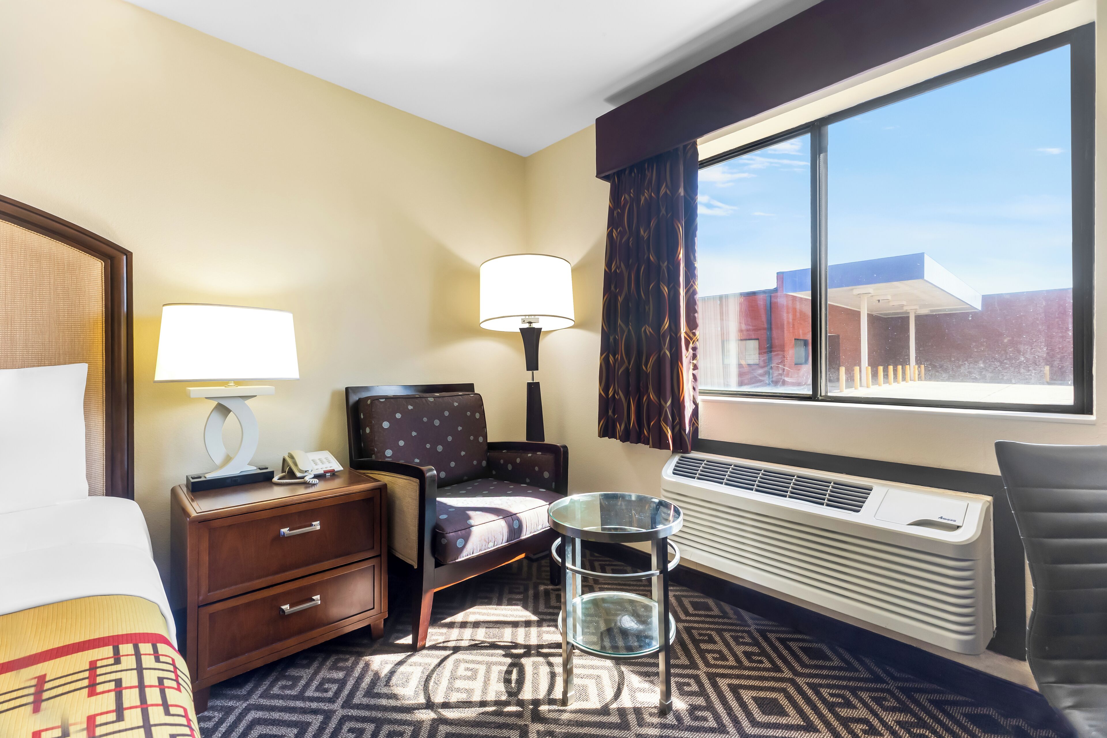 A hotel room with a bed, a nightstand with a lamp, a chair, a small table, and a large window offering a view of buildings outside.