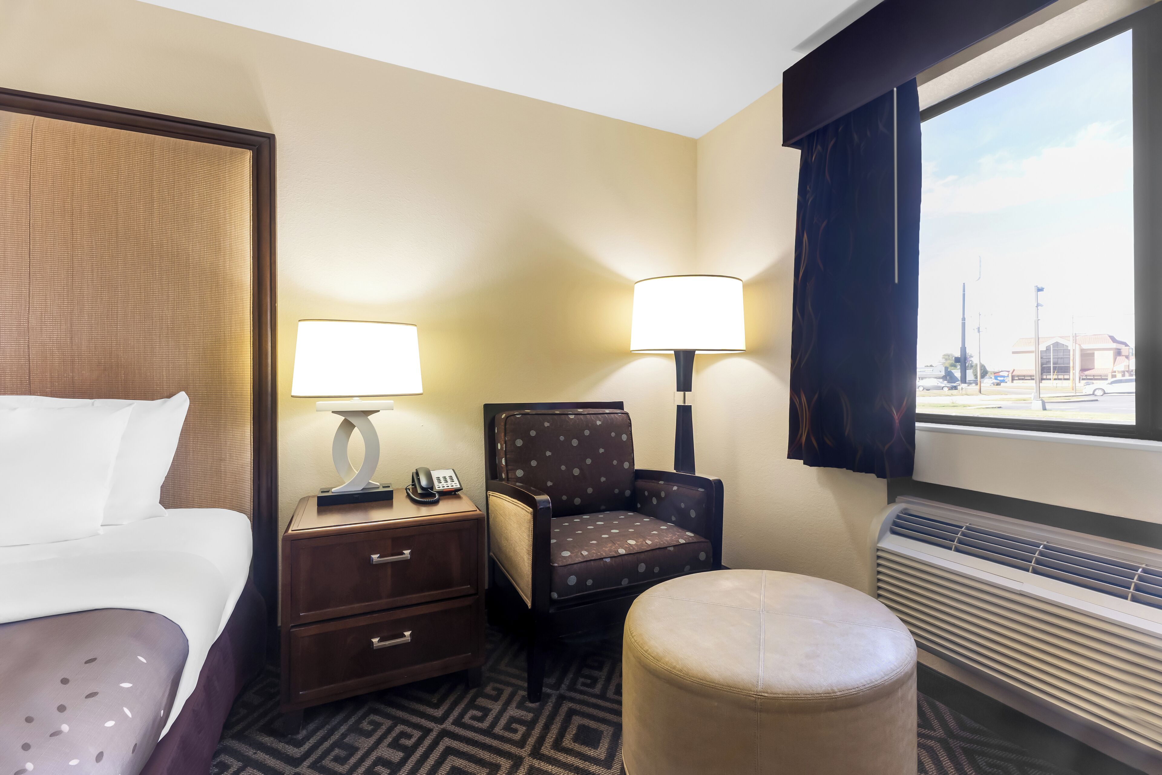 A hotel room with a bed, a chair, a desk with a lamp, a telephone, and a window with a view of the outside.