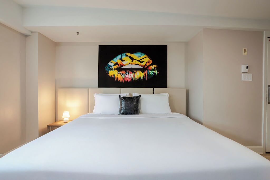 Une chambre d’hôtel avec un grand lit, une tête de lit, une table de chevet avec une lampe et une œuvre d’art représentant des lèvres, accrochée au mur.
