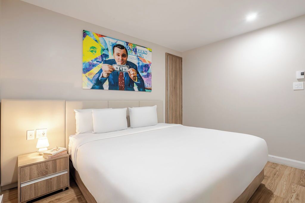 A hotel room with a large bed, a nightstand with a lamp, and a colorful painting on the wall.