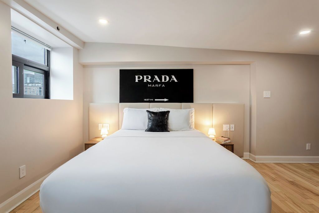 Une chambre d’hôtel avec un grand lit, deux tables de chevet avec des lampes, une fenêtre donnant sur la ville et une enseigne au-dessus du lit sur laquelle on peut lire « PRADA MARFA ».