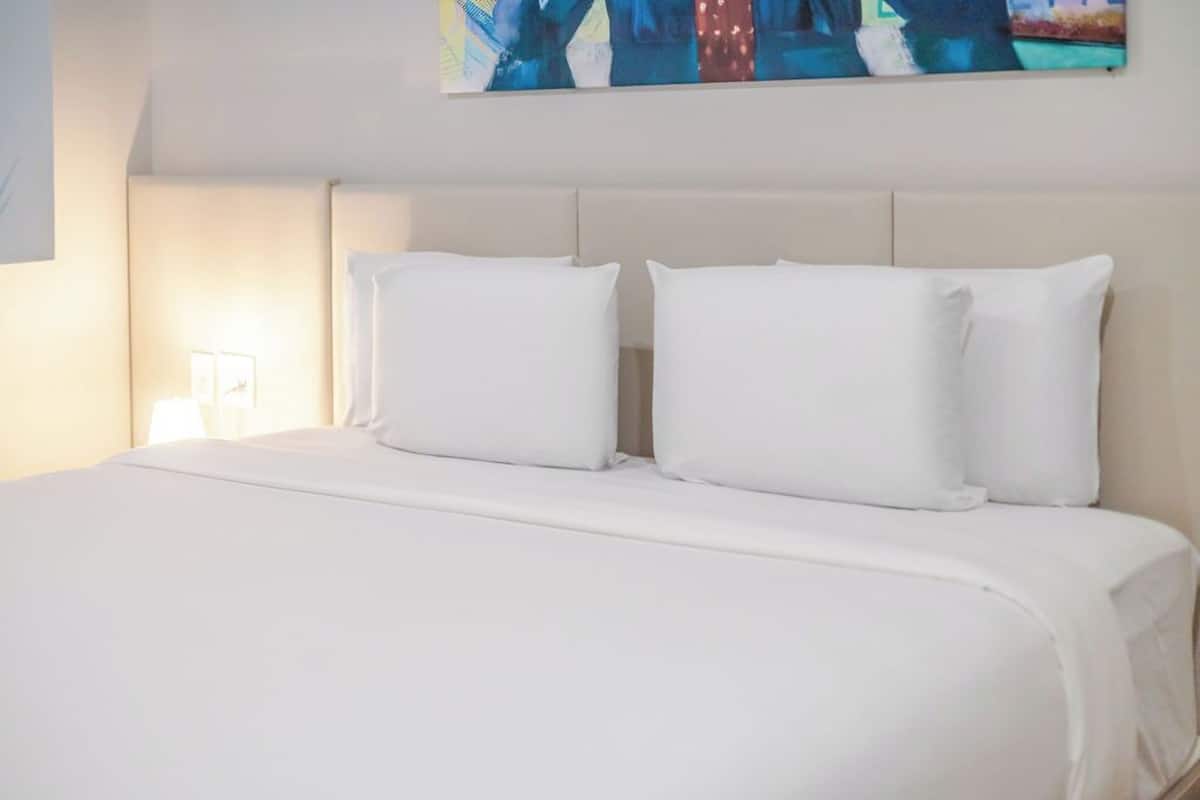 A neatly made bed with white bedding and pillows, a bedside lamp, and a wall with abstract art.