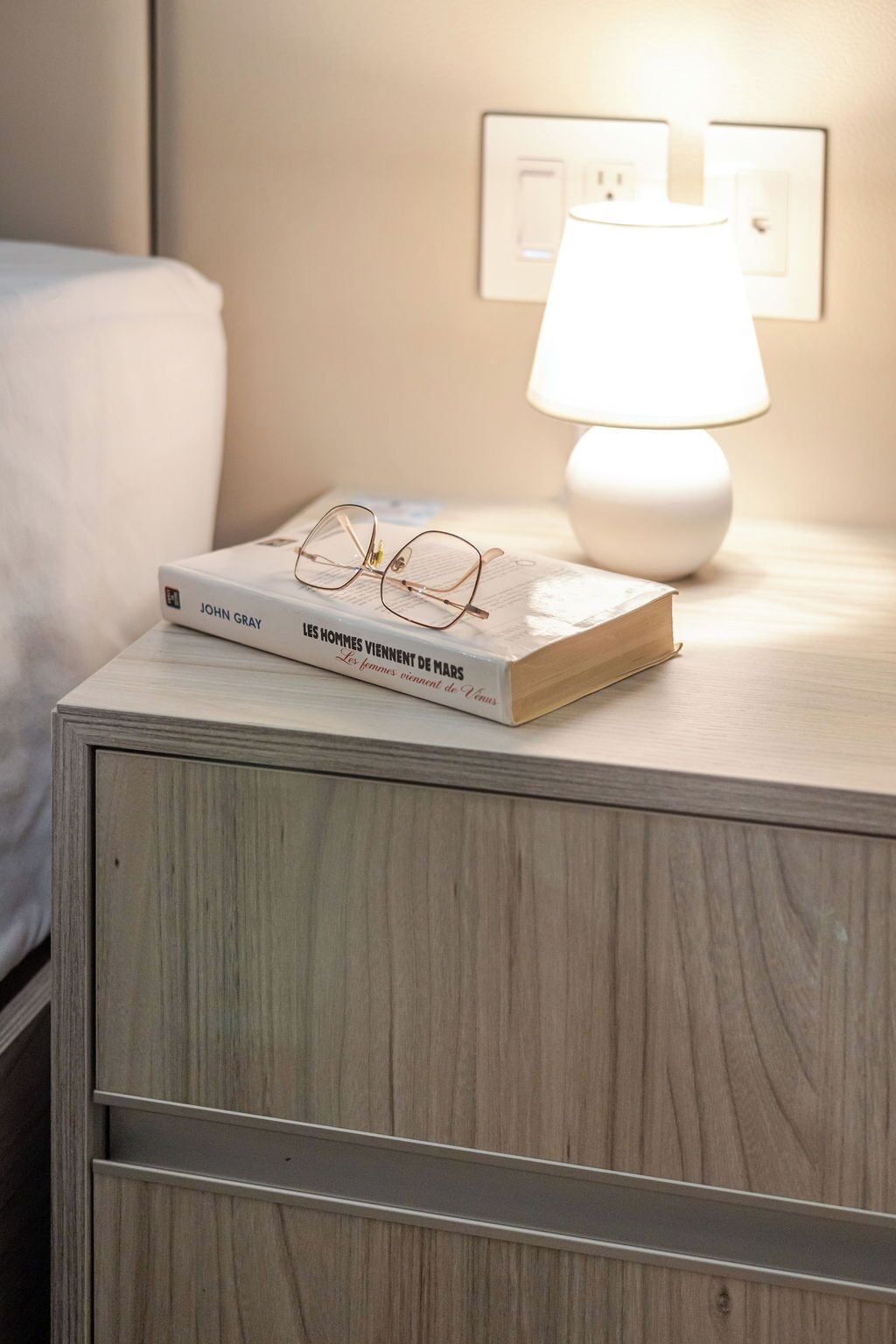 Uma mesa de cabeceira com um livro intitulado 'John Gray' e um par de óculos, uma luminária branca e uma tomada na parede.