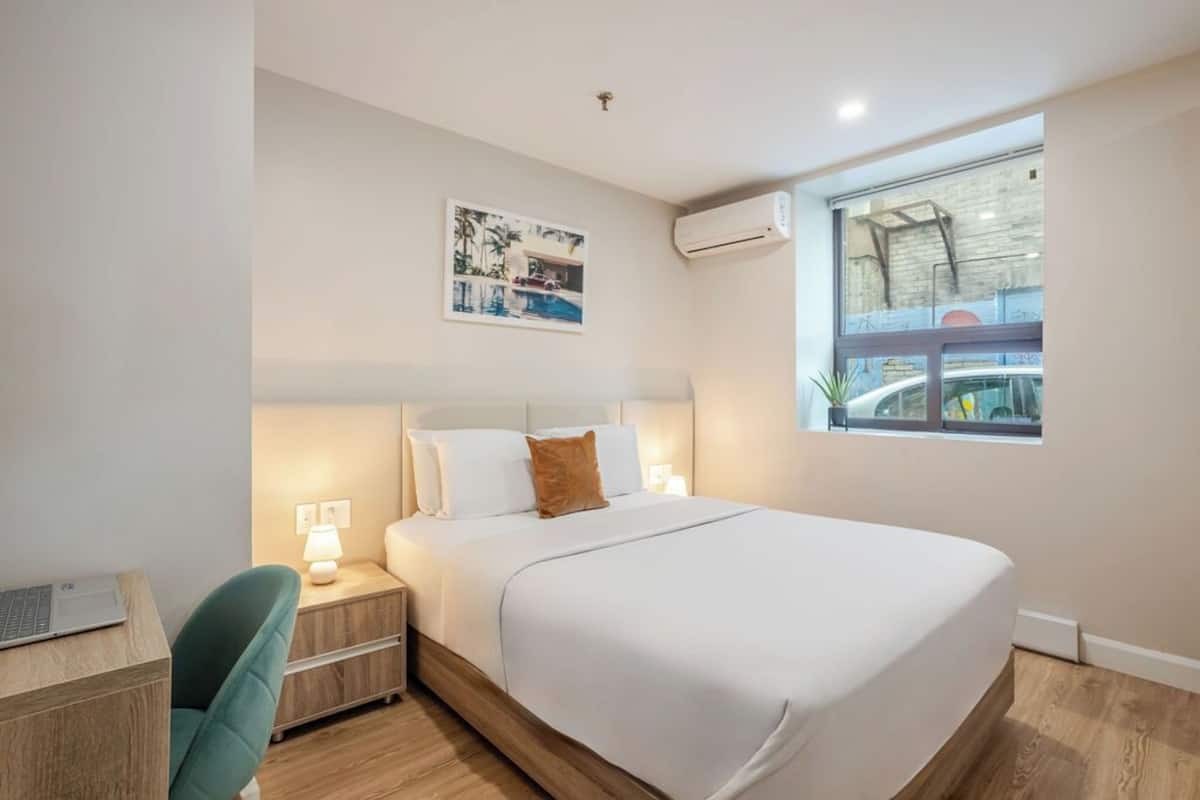 A modern hotel room with a large bed, a desk with a laptop, a chair, a nightstand with a lamp, and a window with a view of a building.