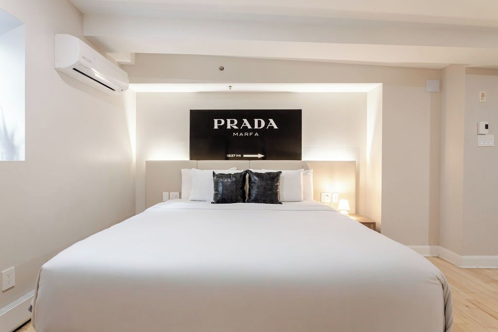 A modern hotel room with a large bed, a mounted TV displaying the Prada logo, and a wall-mounted air conditioning unit.