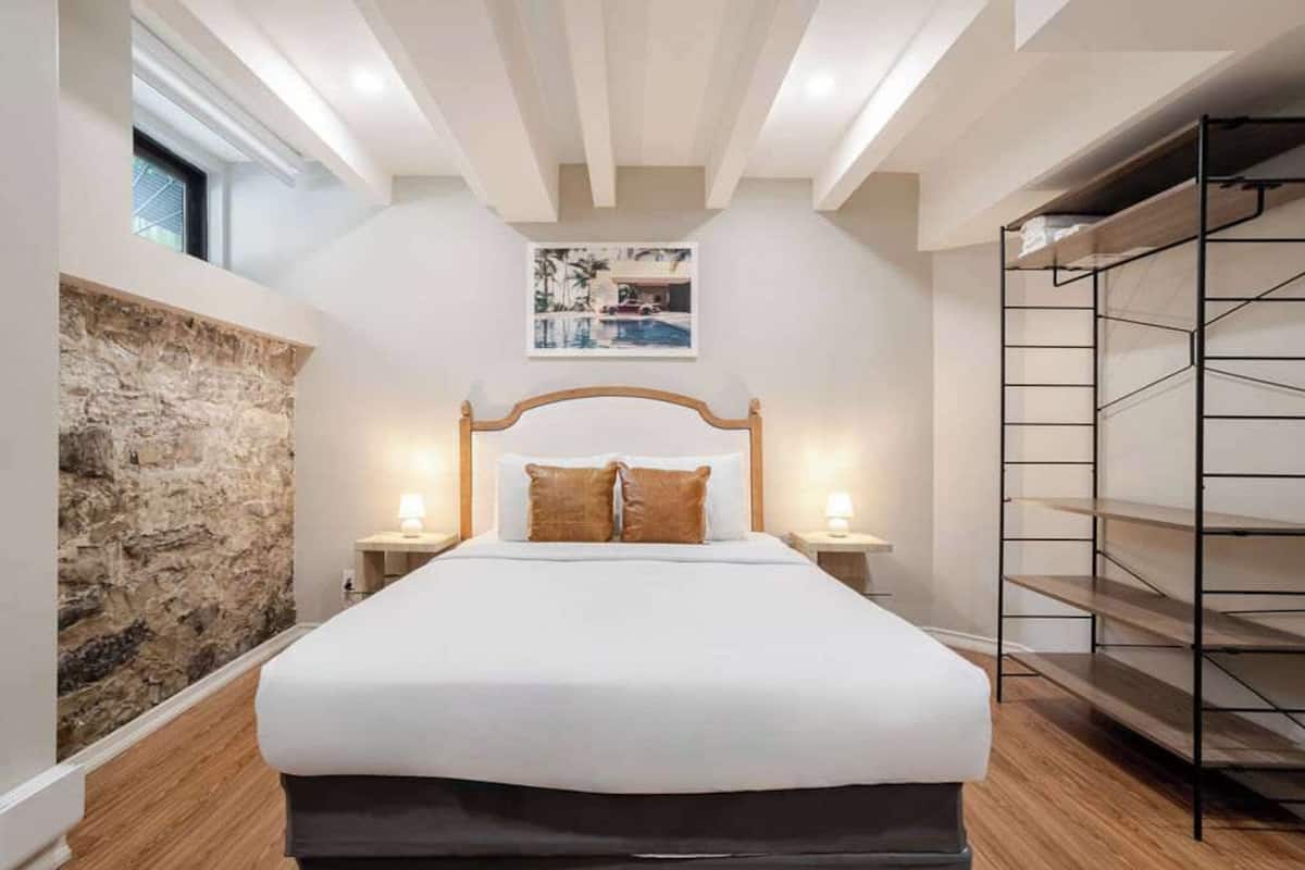 A bedroom with a large bed, stone wall, wooden headboard, two bedside lamps, and a framed picture on the wall.