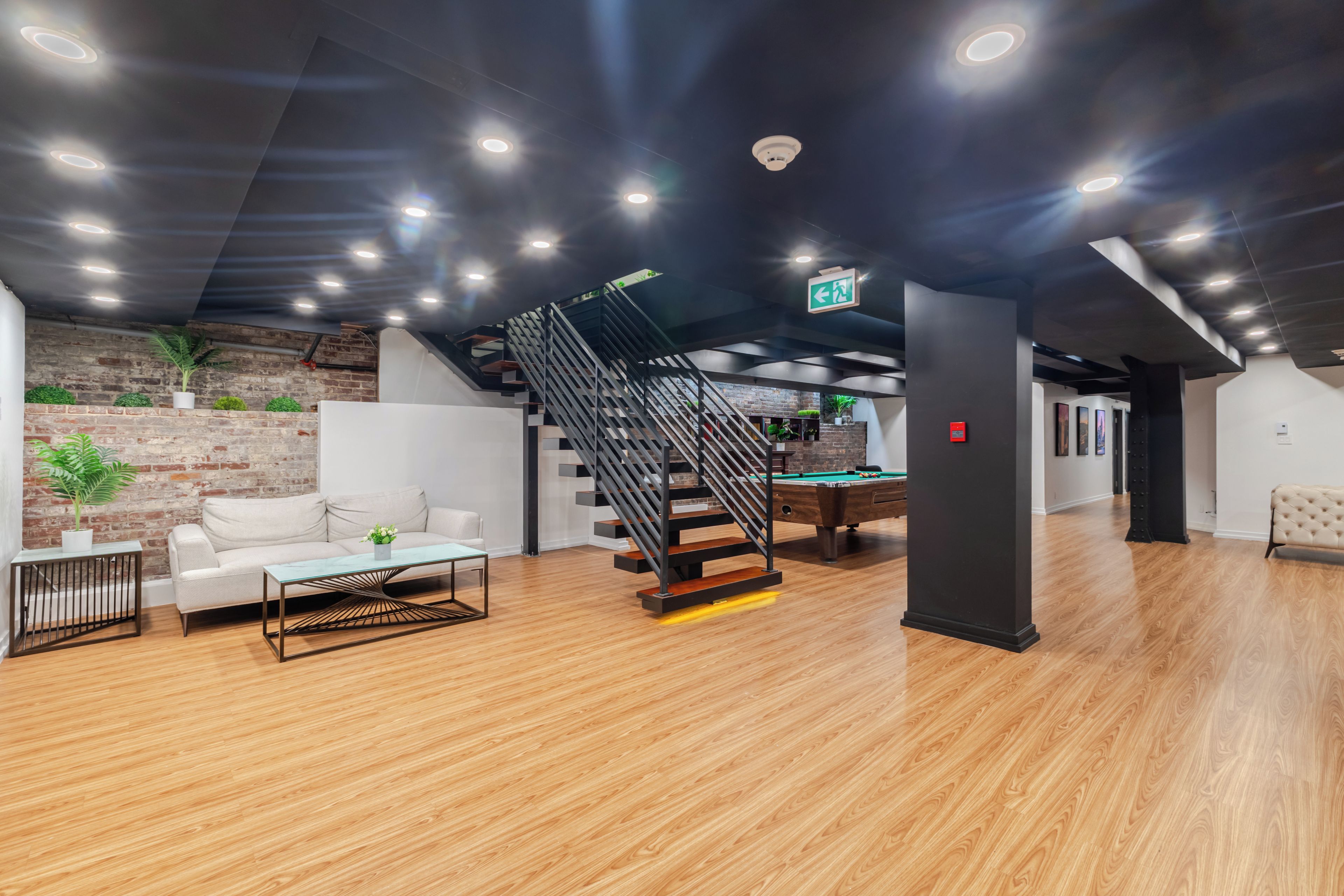 A modern indoor space with a wooden floor, a white sofa, a glass-top coffee table, a pool table, and a black staircase.