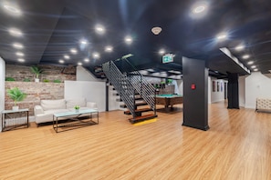 A modern indoor space with a wooden floor, a white sofa, a glass-top coffee table, a pool table, and a black staircase. - Olympia Hotel By Lodgo (Montreal)