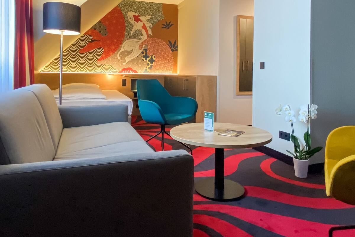 A hotel room with a sofa, a round table with a sign, a blue chair, a yellow chair, and a decorative wall with a fish pattern.