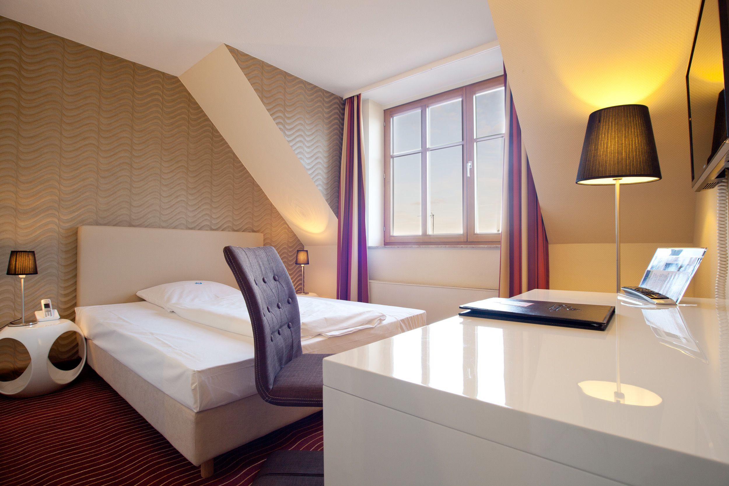 A hotel room with a bed, a desk, a chair, a lamp, and a window.