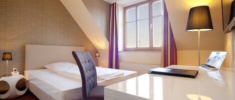 A hotel room with a bed, a desk, a chair, a lamp, and a window.