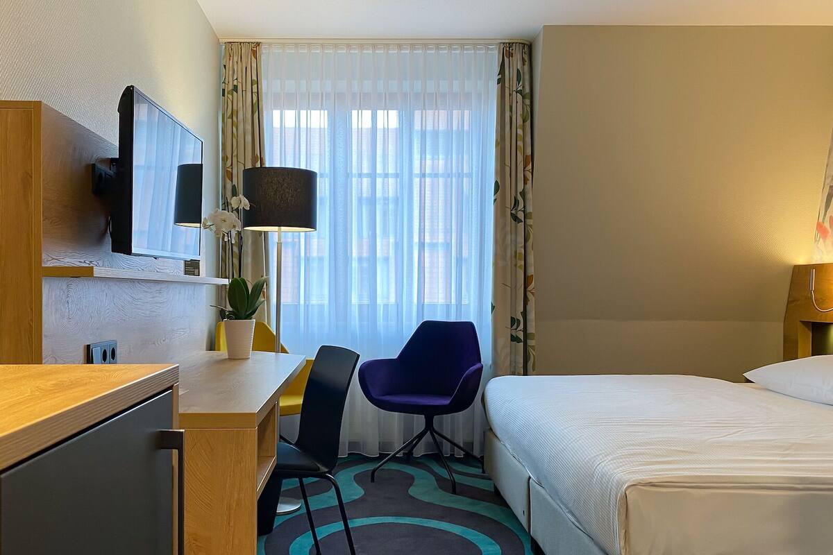 A hotel room with a bed, a desk with a chair, a TV, a lamp, and a window with curtains.