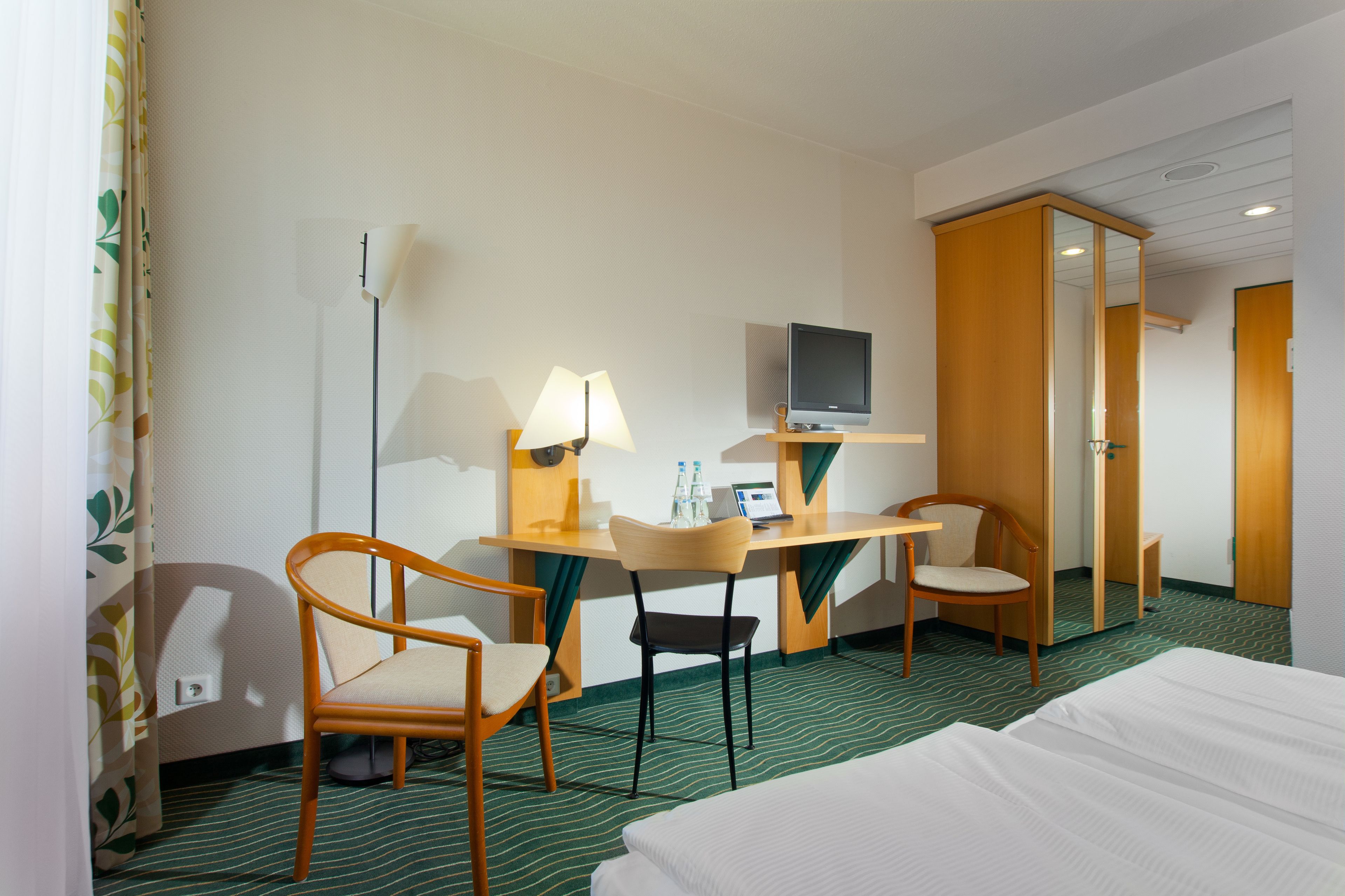 Ein Hotelzimmer mit einem Bett, zwei Sesseln, einem Schreibtisch mit Computer und Lampe, einem Fernseher und einem Kleiderschrank.