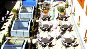 Un patio con mobiliario exterior, una zona techada y un edificio con tejados rojos y numerosas ventanas.