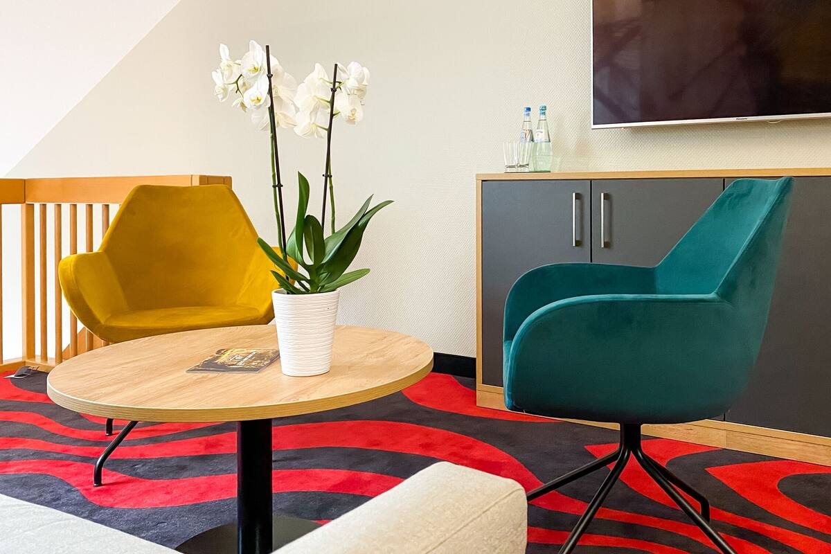 A modern room with a round wooden table, a yellow chair, a teal armchair, a white potted plant, a grey cabinet, and a flat-screen TV on the wall.