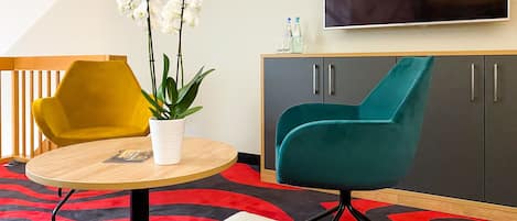 A modern room with a round wooden table, a yellow chair, a teal armchair, a white potted plant, a grey cabinet, and a flat-screen TV on the wall.