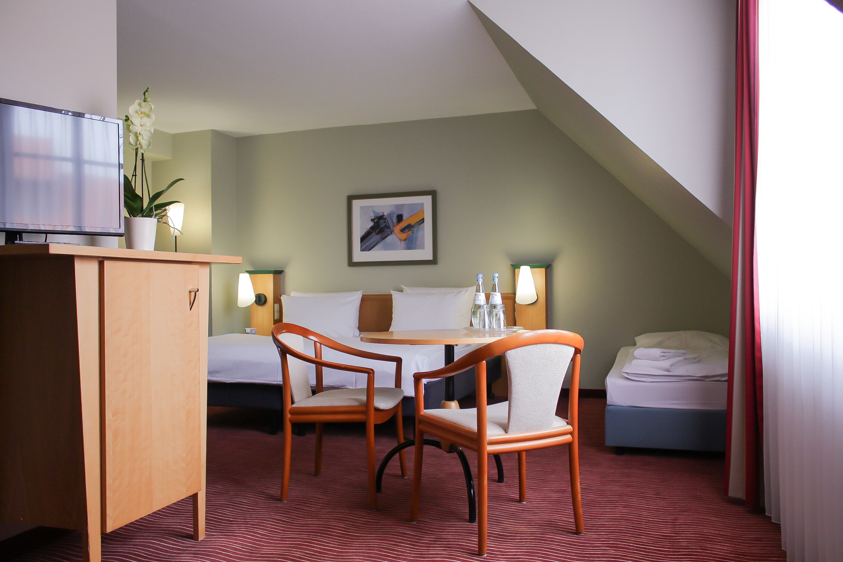 Ein Hotelzimmer mit Bett, Schreibtisch mit Fernseher, zwei Stühlen, Tisch und einer Zimmerpflanze.