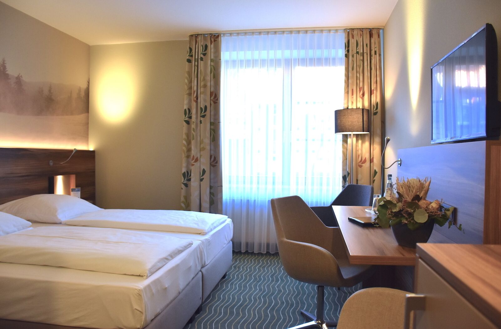 Habitació d'hotel amb un llit, una taula de treball amb una cadira, una làmpada, un televisor i una finestra amb cortines.