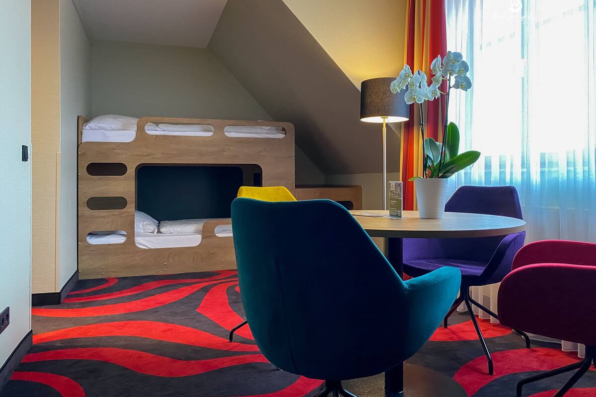 A hotel room with a bunk bed, a desk with a lamp, a potted plant, and colorful chairs.