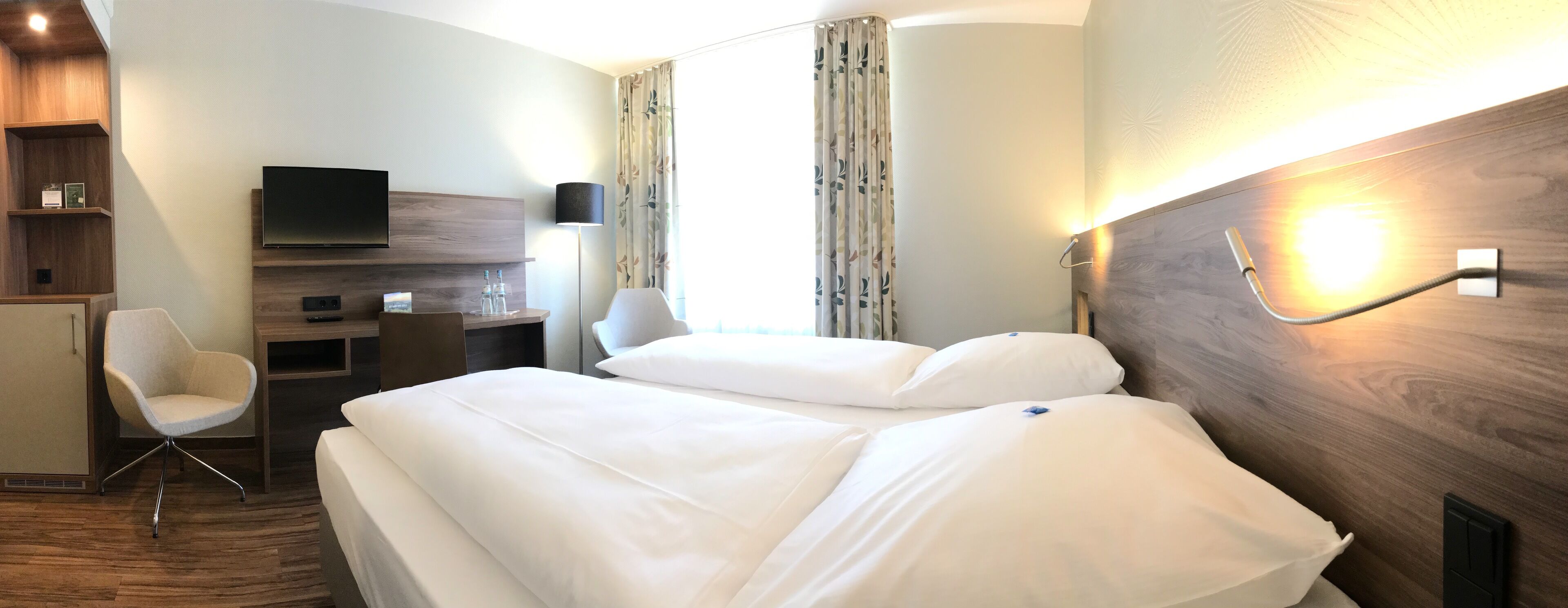 A hotel room with a large bed, a desk with a chair, a flat-screen TV, and a lamp.