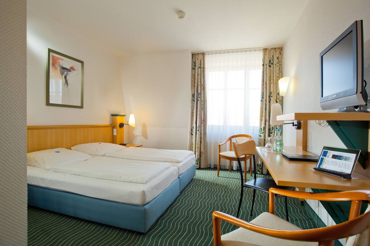 A hotel room with two beds, a desk with a laptop, a chair, a TV, and a window with curtains.
