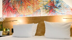 Habitación de hotel con cabecera de madera, dos almohadas blancas, mesita de noche con un libro y una planta pequeña, y un cuadro abstracto colorido sobre la cama.