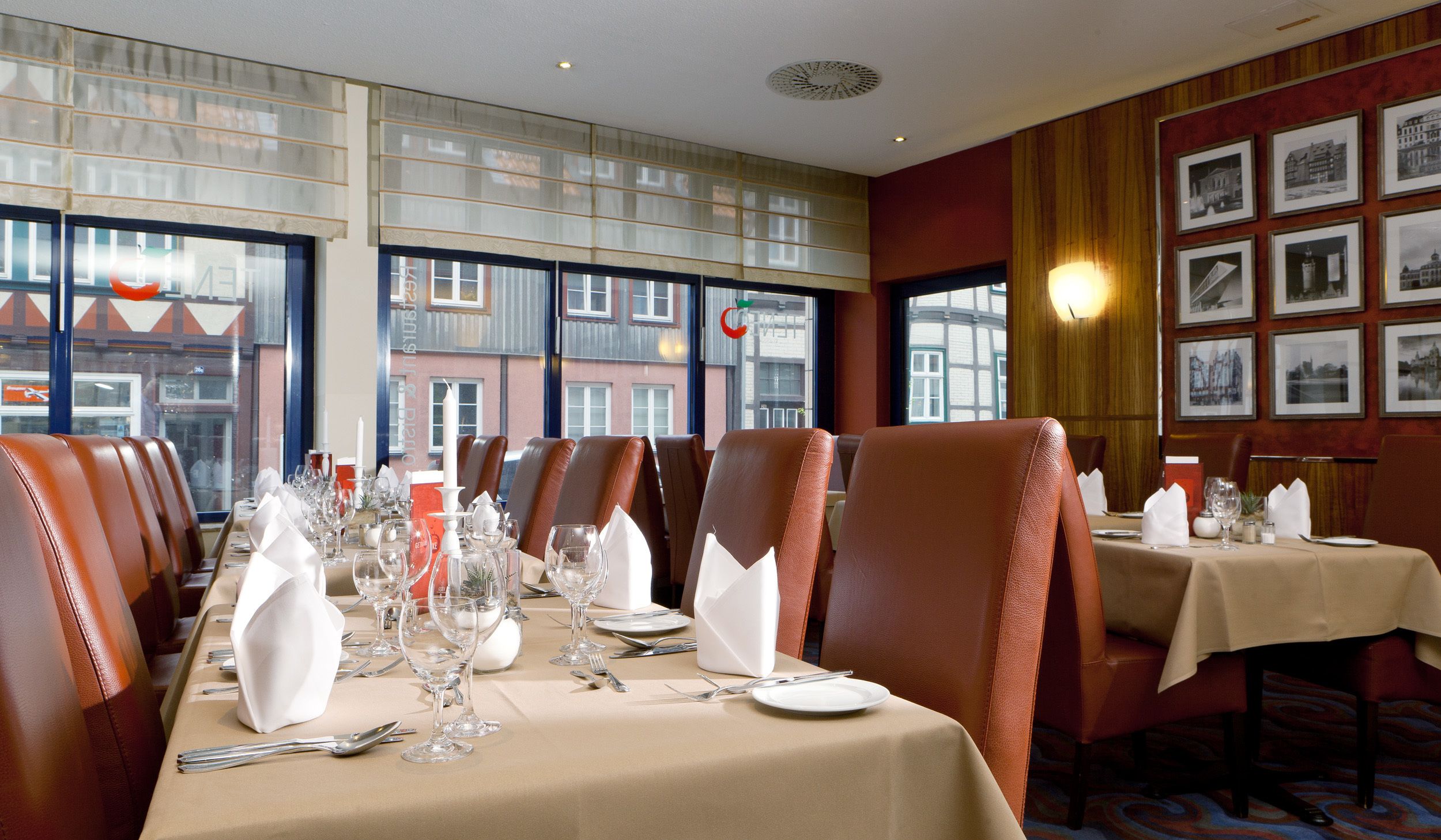 Un restaurant avec de longues tables dressées pour le repas, des fauteuils en cuir et des tableaux encadrés aux murs.