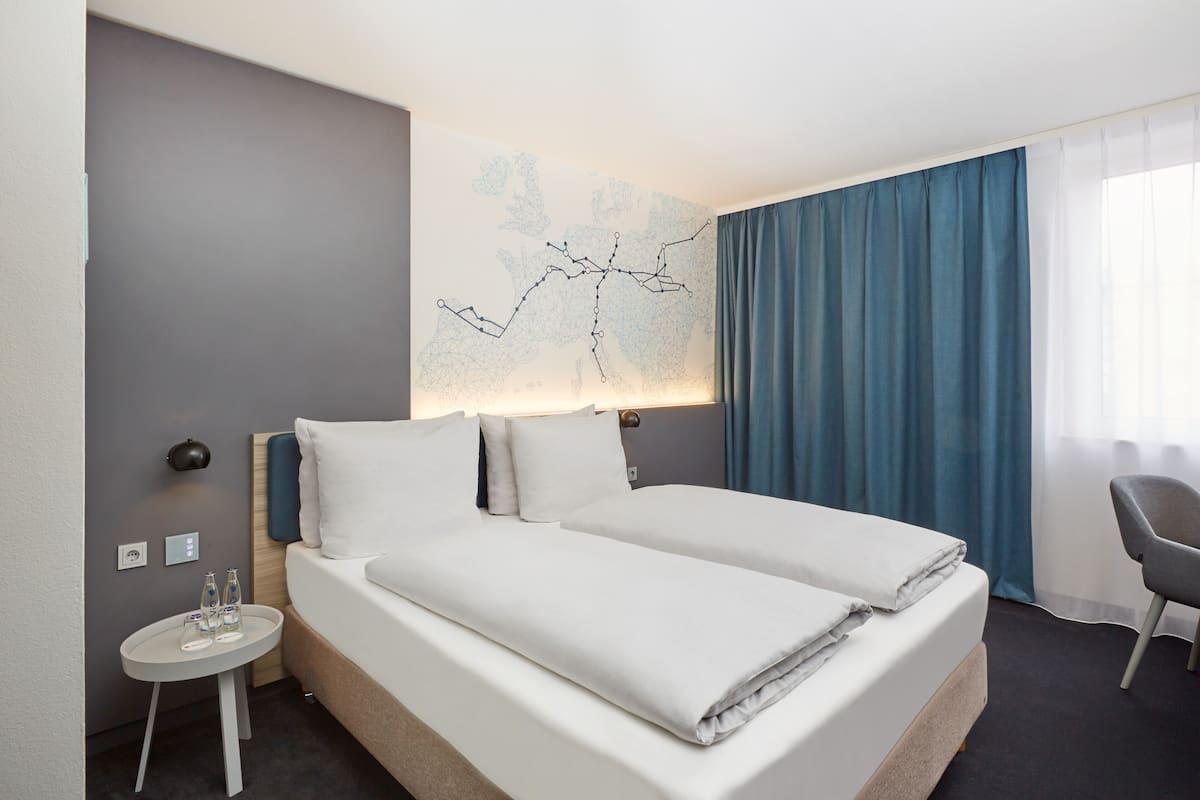 A hotel room with a bed, a nightstand, a lamp, and a window with curtains.