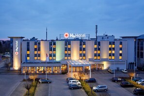 A modern hotel with a prominent H4Hotel sign, a covered entrance, and a parking lot with several cars. - H4 Hotel Leipzig (Leipzig)