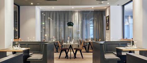 A modern dining area with high tables and booths, large windows with curtains, and recessed ceiling lighting.