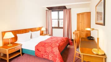 A hotel room with a bed, two bedside tables with lamps, a desk with a chair, a wooden wardrobe, and a window with curtains.
