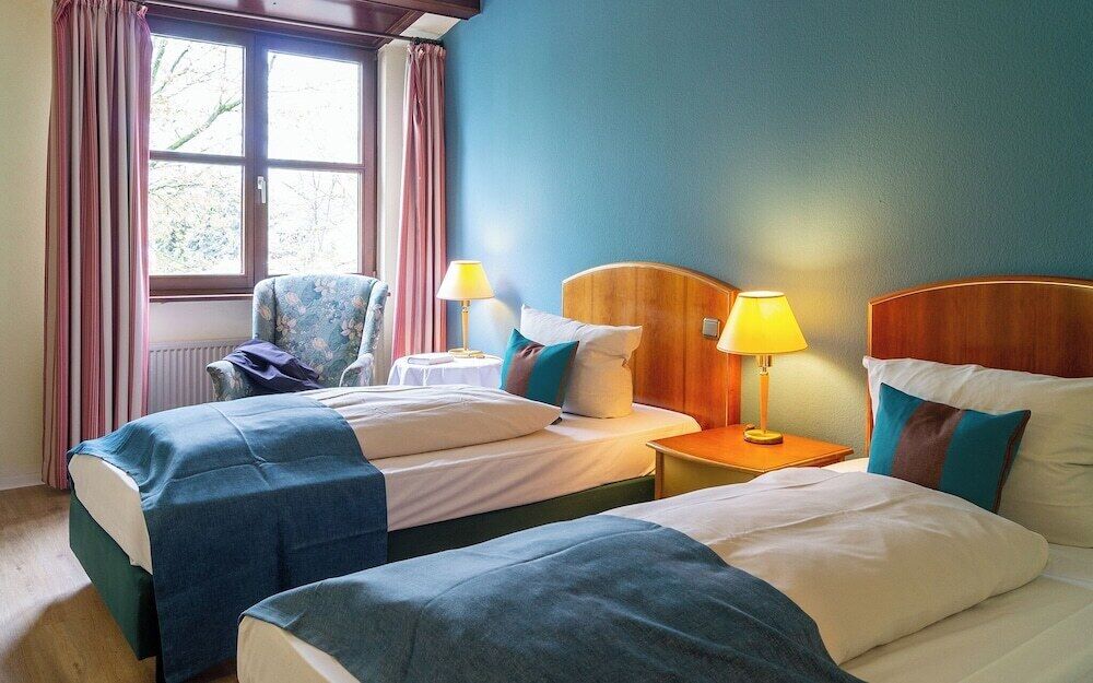A hotel room with two beds, a chair, a window with curtains, and two bedside lamps.