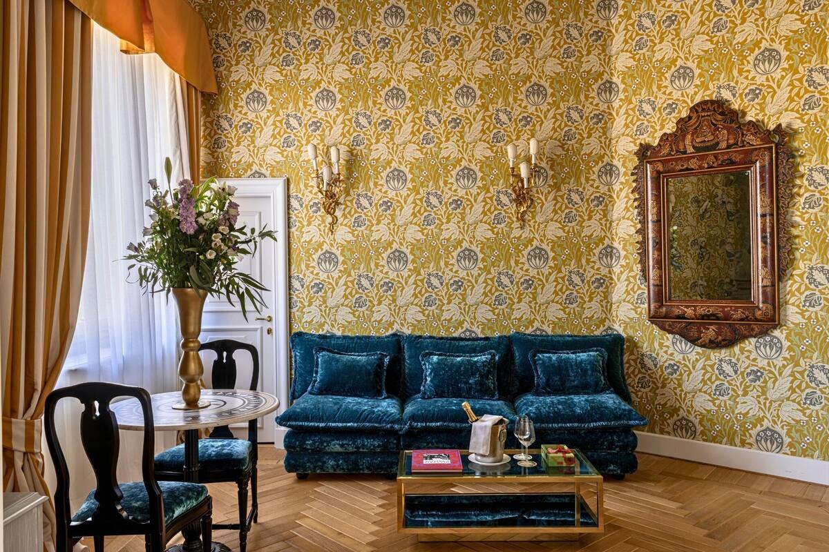 A room with a blue velvet sofa, a round table with chairs, a vase with flowers, and patterned wallpaper.