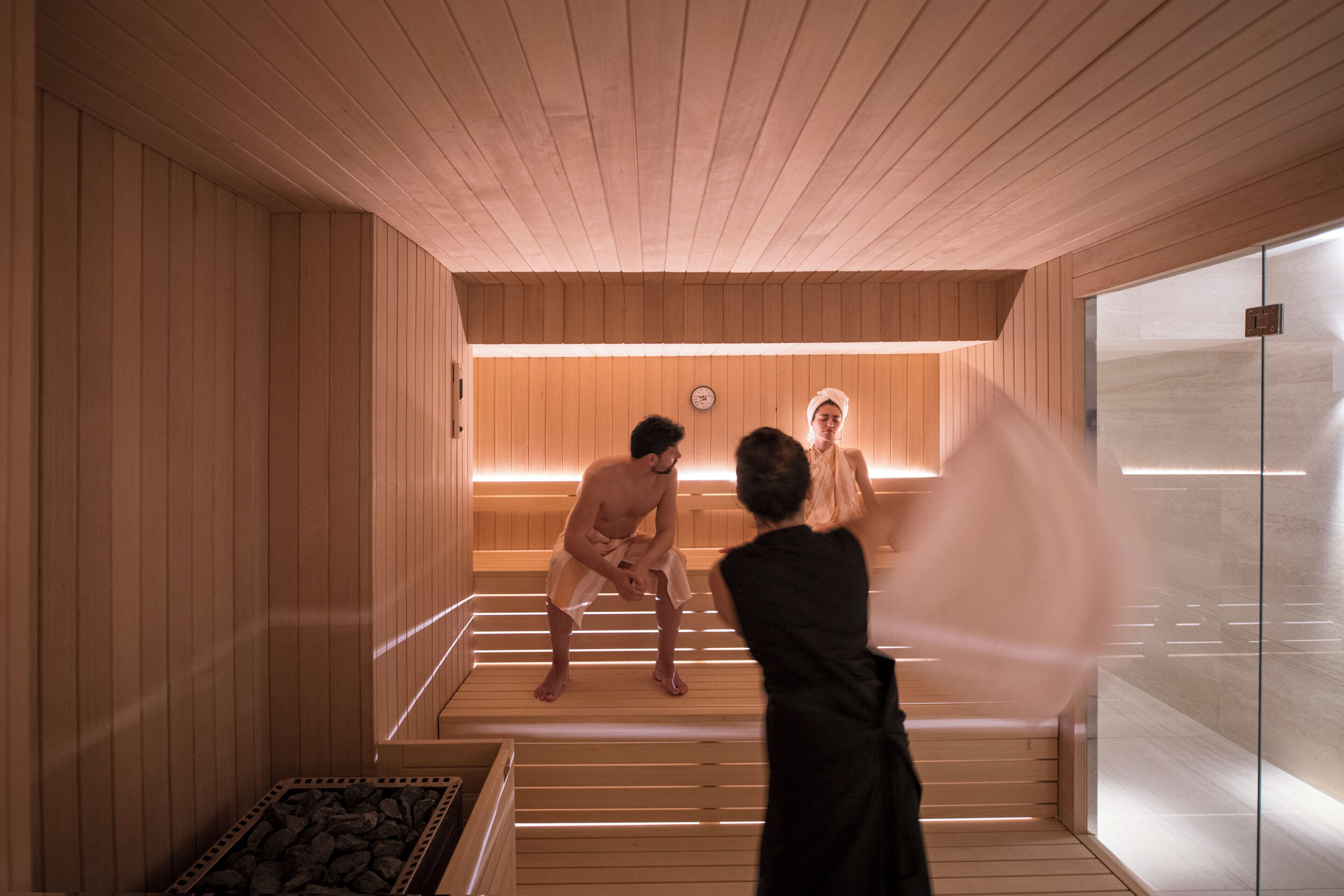 Un sauna avec des murs et des bancs en bois, une horloge au mur et une personne prenant une photo.