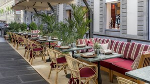 Un café al aire libre con sillas de mimbre y cojines rojos, mesas con sombrillas y plantas en macetas.