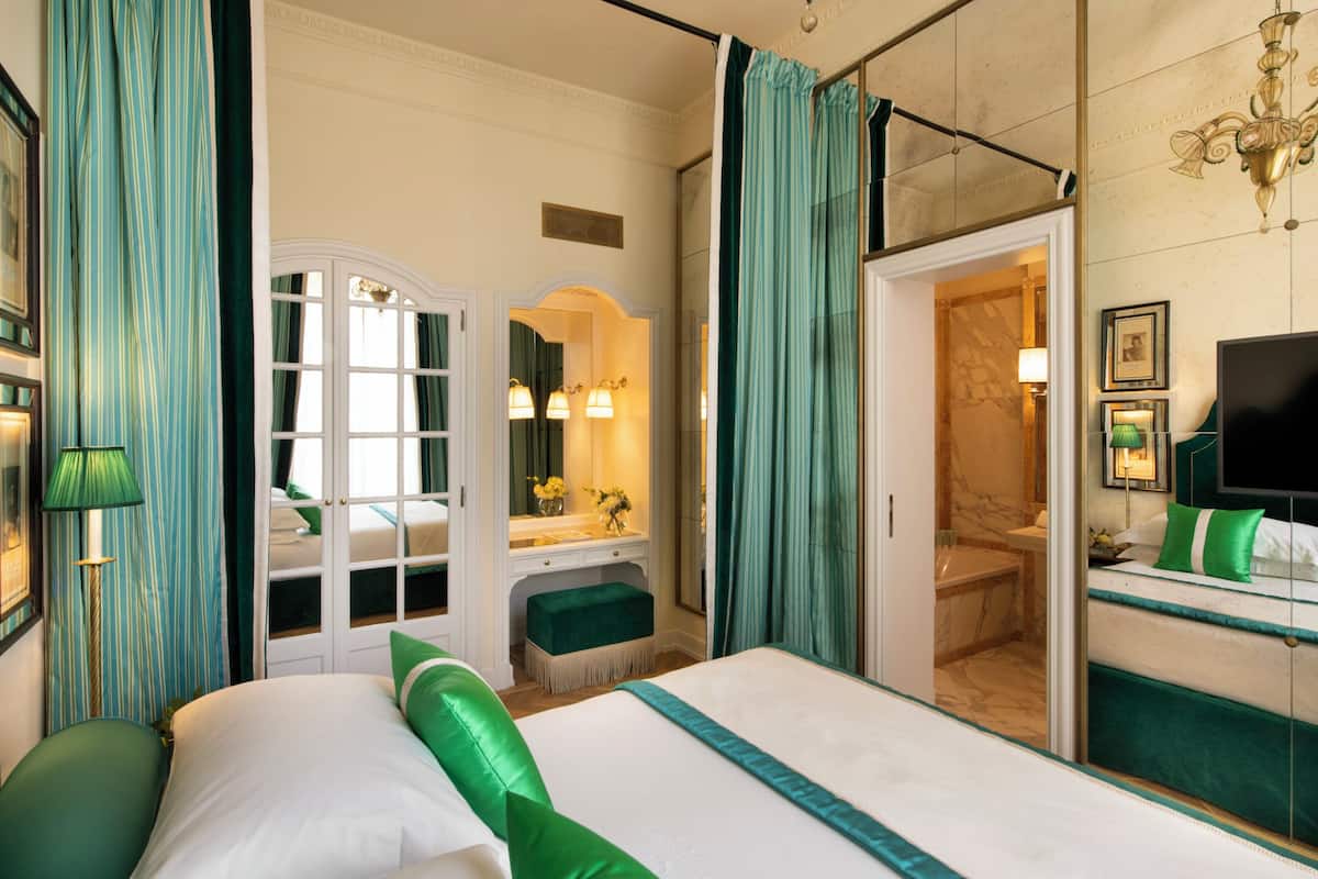Habitación de hotel con una cama grande, una tocador con espejo, un banco tapizado en verde y un baño visible a través de una puerta abierta.