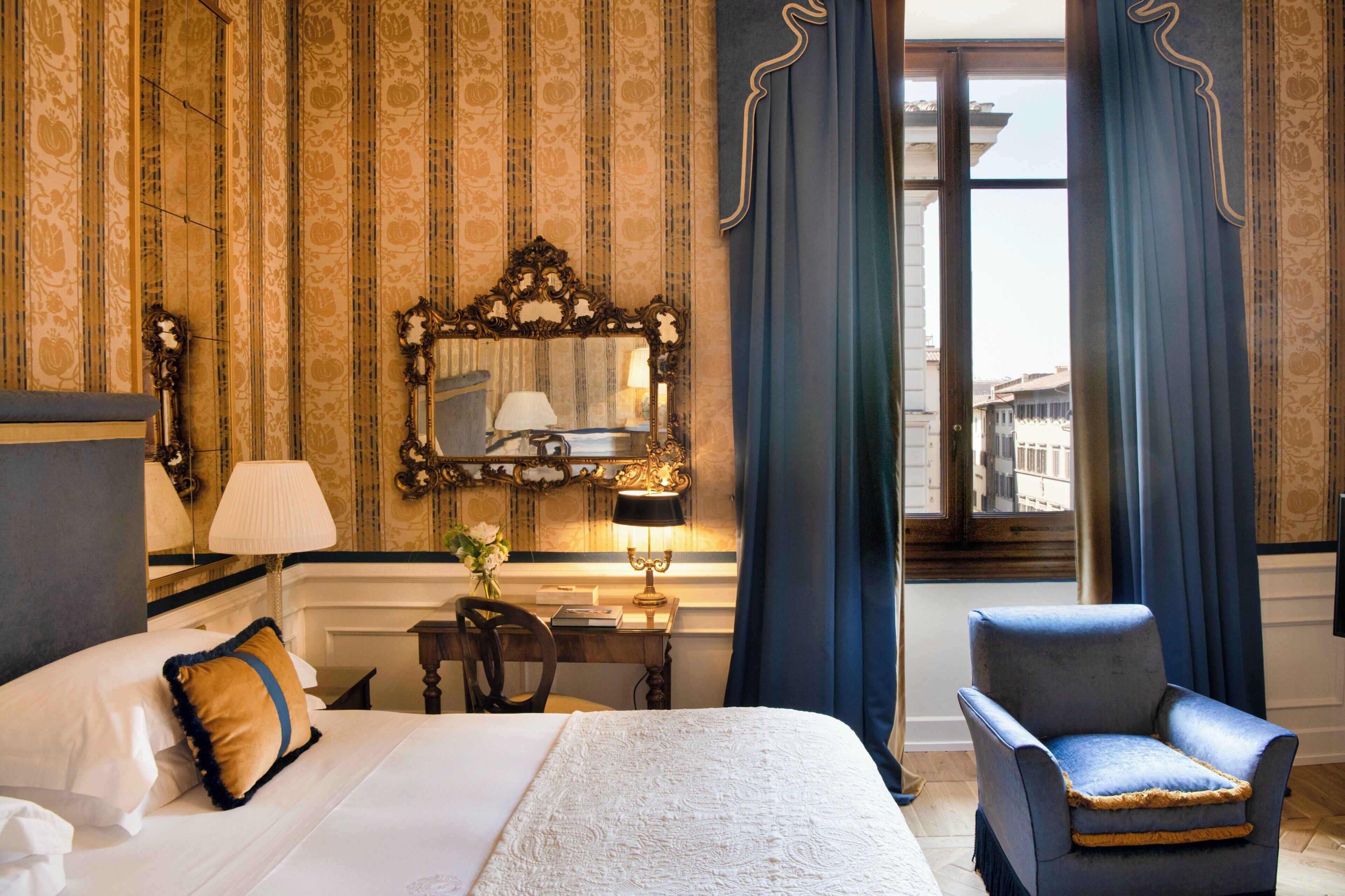 A hotel room with a large bed, a chair, a desk with a lamp, a mirror, and a view of the city through a window.