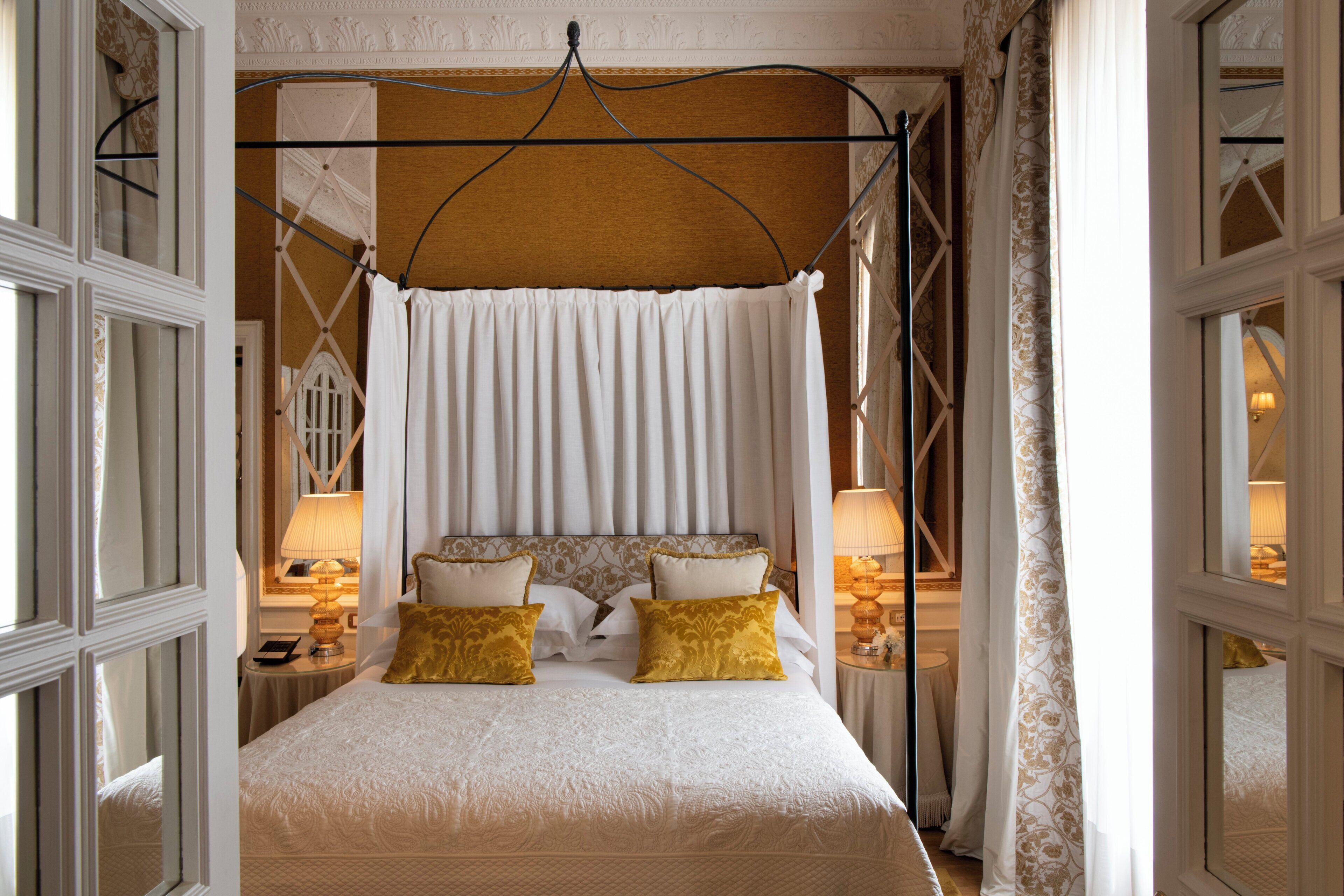 A bedroom with a canopy bed, two bedside tables with lamps, and a view of the room through a window.