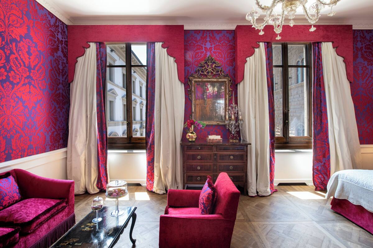 A room with a red sofa, a wooden dresser, and a bed. There are two windows with curtains, and a chandelier hangs from the ceiling.