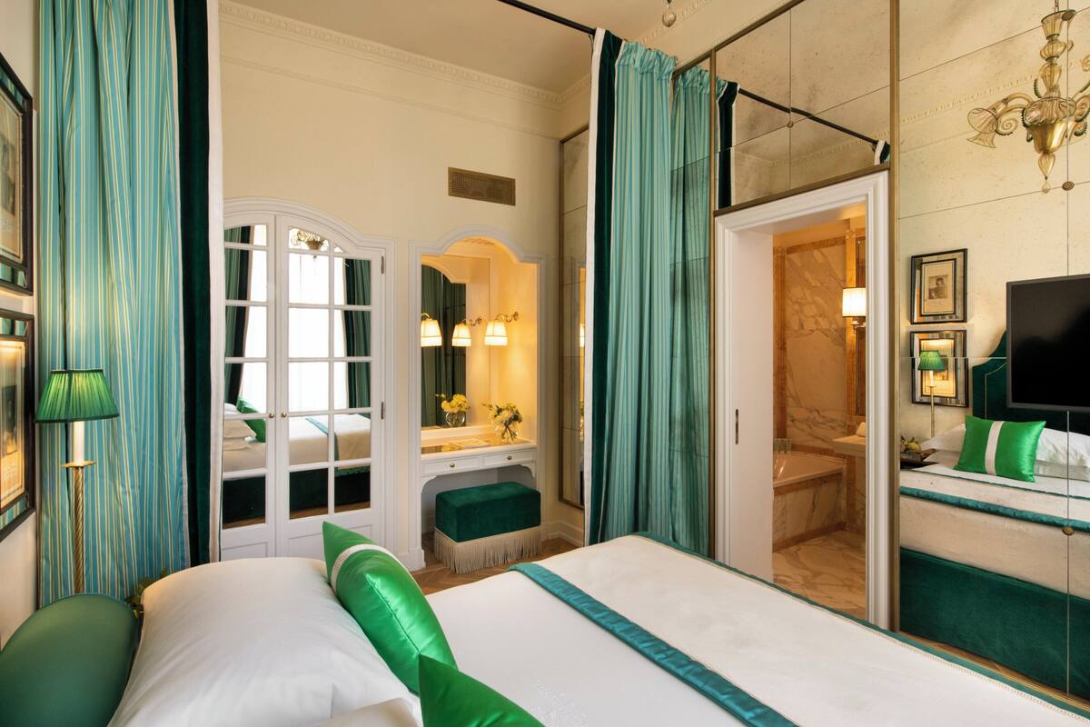 A hotel room with a large bed, a vanity with a mirror, a green upholstered bench, and a bathroom visible through an open door.