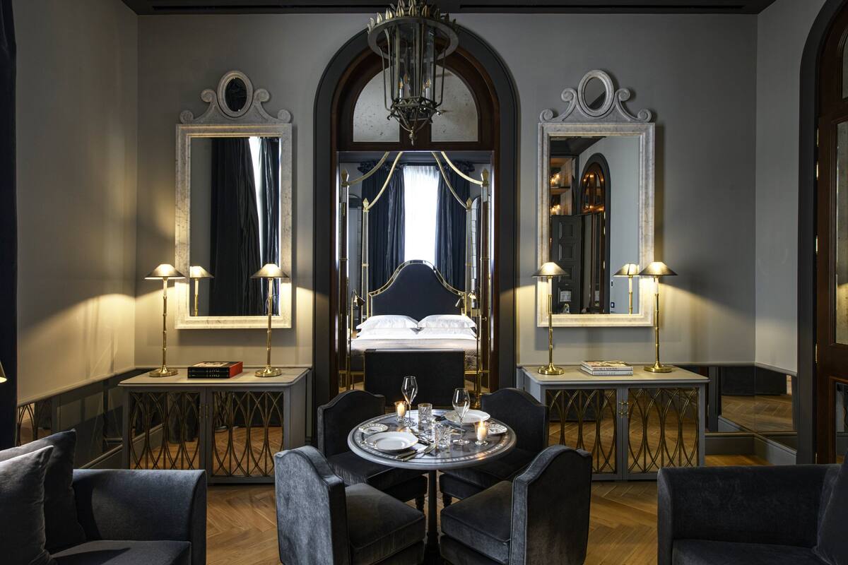A luxurious bedroom with a large bed, two bedside tables with lamps, a round dining table with chairs, and a large mirror above the bed.