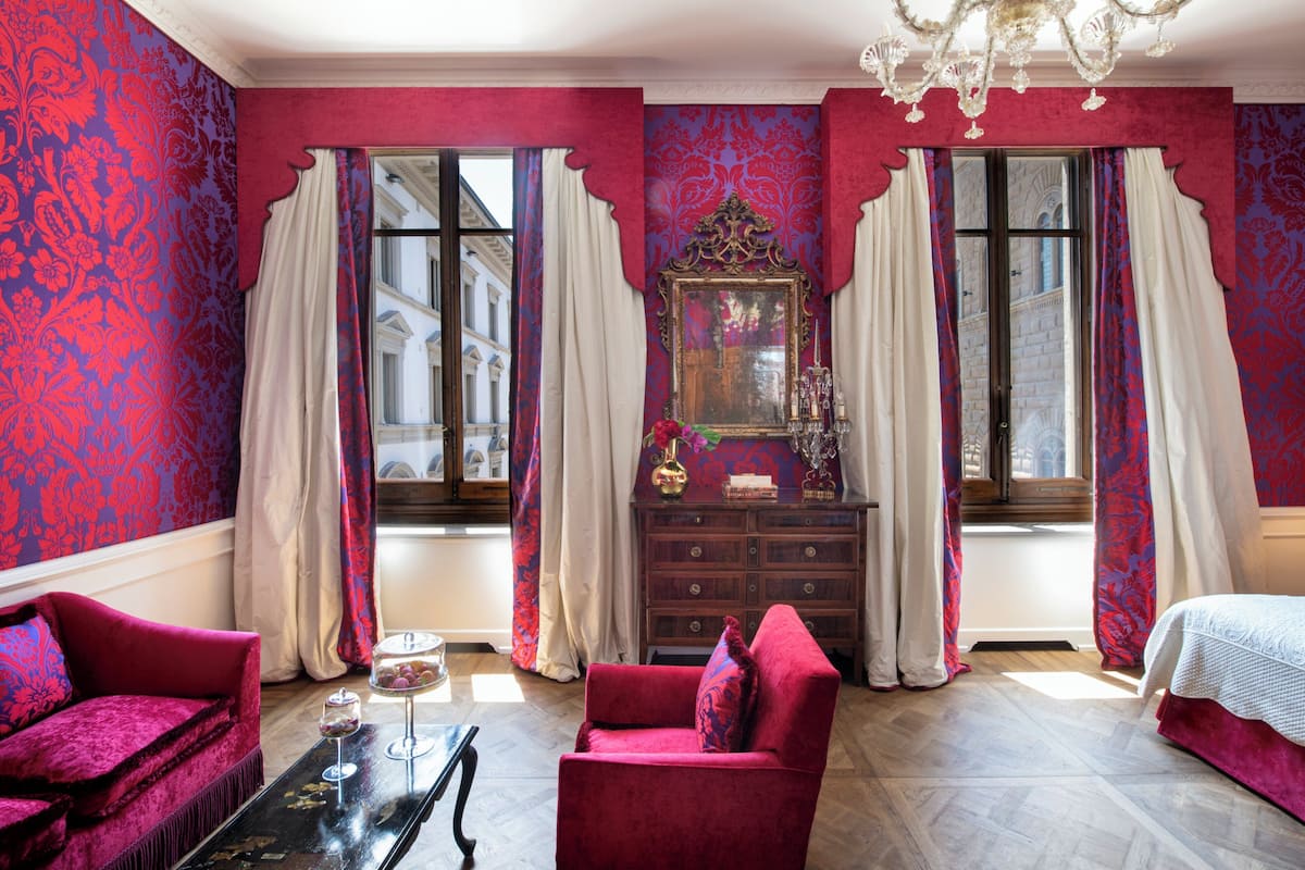Una habitación con un sofá rojo, un aparador de madera y una cama. Hay dos ventanas con cortinas, y un candelabro cuelga del techo.