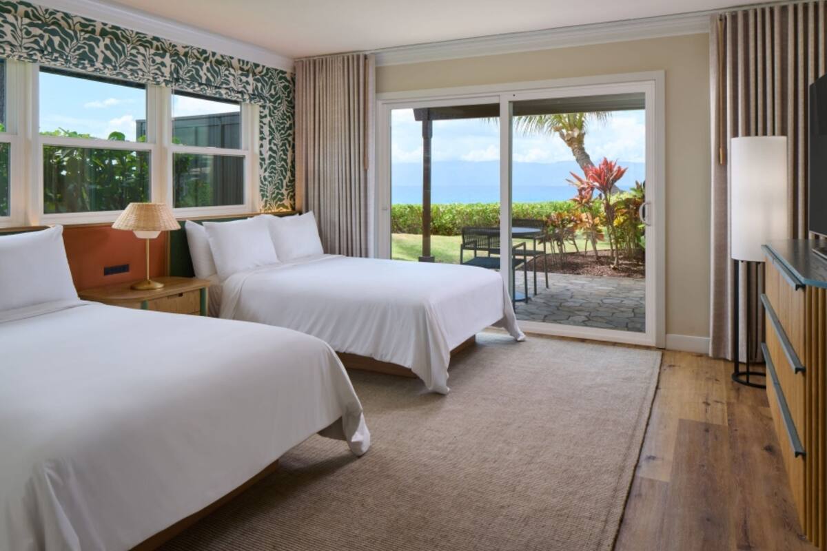 Habitación de hotel con dos camas, un ventanal con vistas al exterior, una puerta corrediza de vidrio que da a un patio y un televisor a la derecha.
