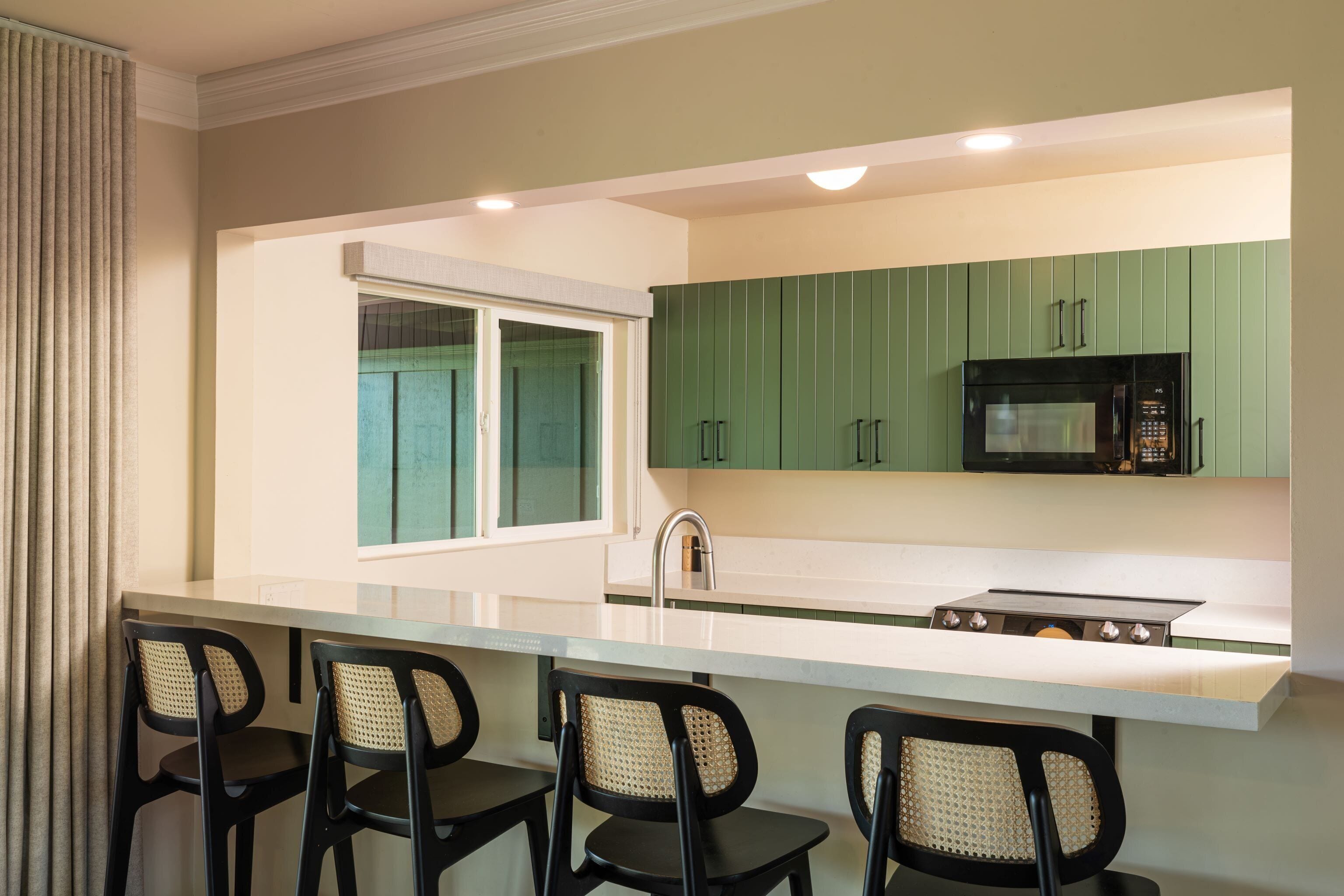 Une cuisine moderne avec des meubles de couleur verte, un four à micro-ondes et une cuisinière à gaz. On y trouve trois chaises noires avec des assises en osier tressé.