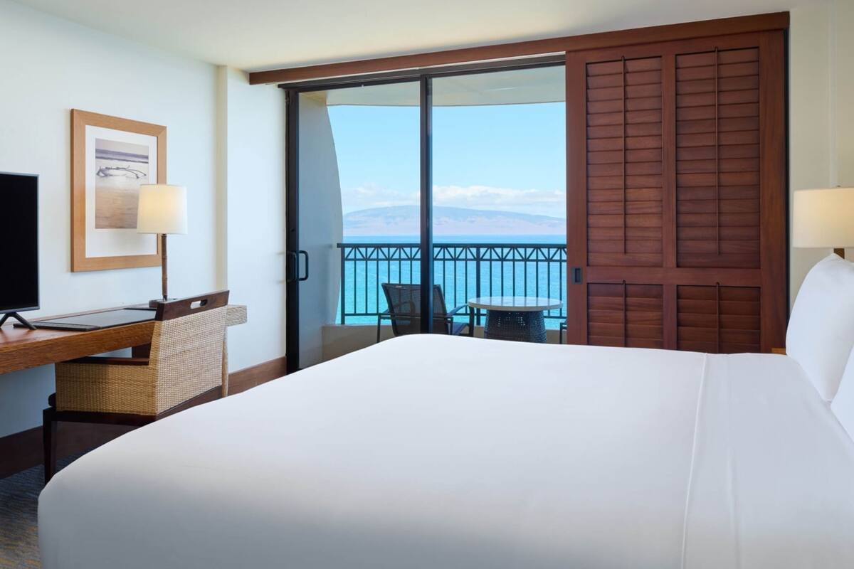 Habitación de hotel con una cama grande, un escritorio con computadora, una silla, una mesita y un balcón con vista al mar.