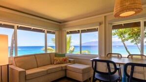 A room with a sofa, dining table, chairs, and large windows offering a view of the ocean.