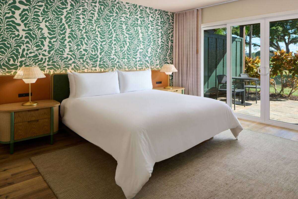 Habitación de hotel con una cama grande, dos mesitas de noche con lámparas, un papel tapiz estampado y un ventanal corredero que da a un espacio exterior.