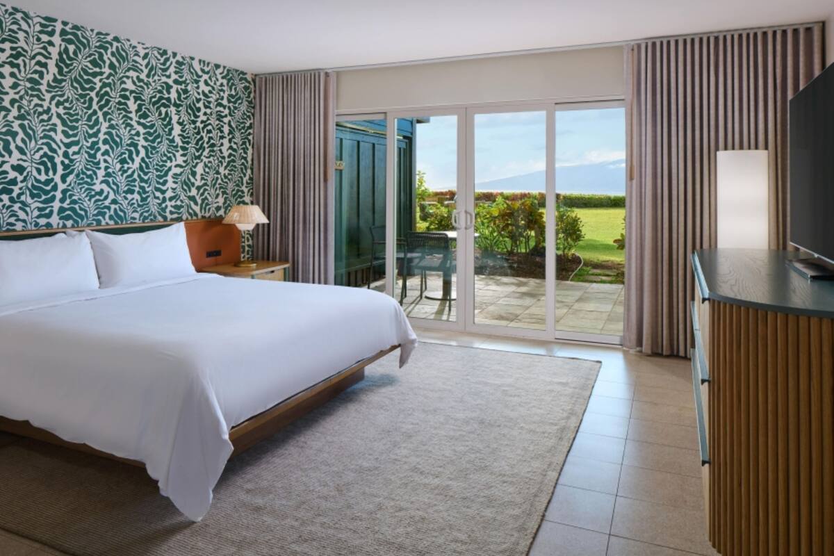 Habitación de hotel con una cama grande, televisión, escritorio y puerta corrediza de vidrio a un patio con vistas al exterior.