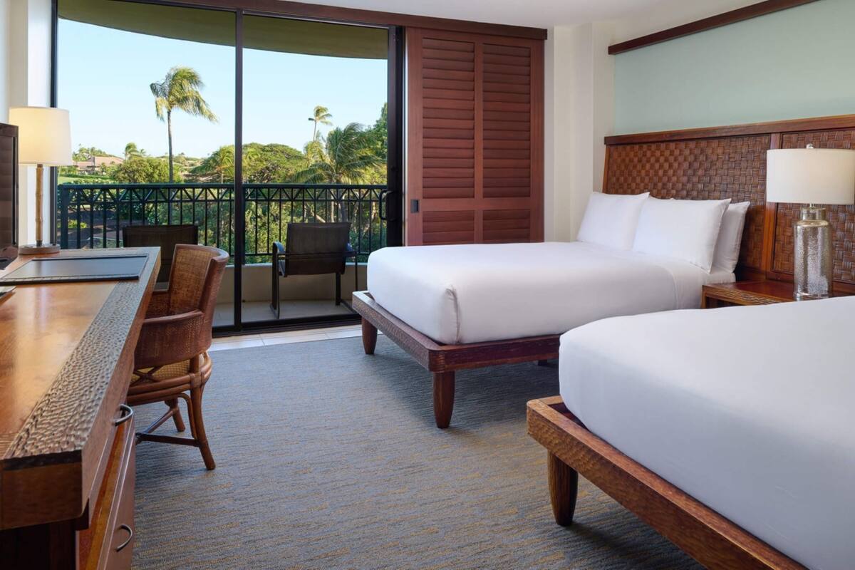 Habitación de hotel con dos camas, un escritorio, una silla y vistas a palmeras a través de una puerta corrediza.
