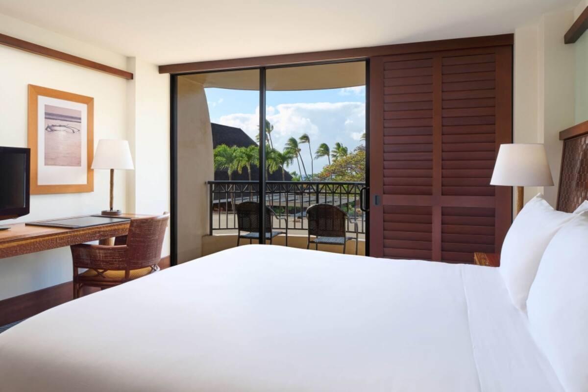 Habitación de hotel con cama, escritorio con computadora, dos sillas, televisión y balcón con vista a palmeras y una montaña.